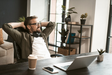 A man is sitting in a chair with his hands on his head and a laptop in front of him. He is wearing glasses and he is relaxed