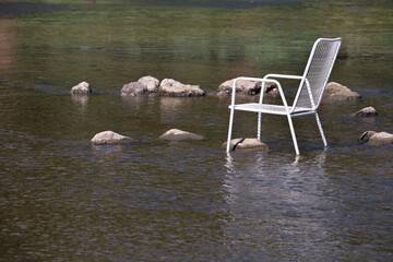 In summer, a white metal chair stands in the middle of a river. There are stones in the middle of...