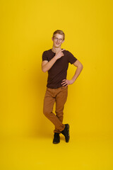 A young man in a brown shirt and tan pants is posing for a photo in front of a yellow background