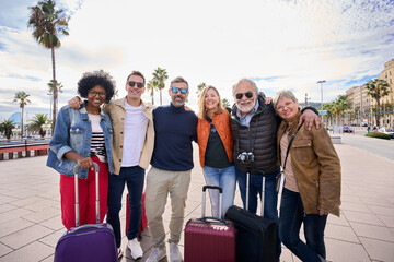 Group of middle aged tourist friends posing embraced and looking camera happy celebrating together their holidays. Diverse tourism people with their luggage in European city street on sunny winter day