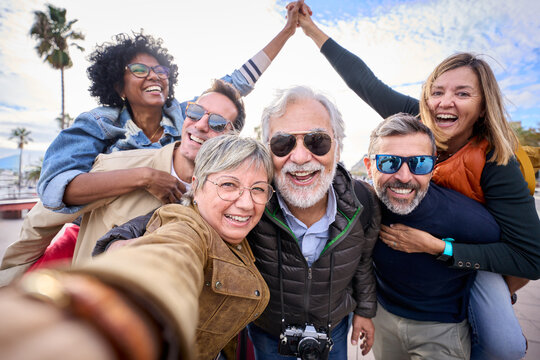 Group diverse funny mature tourist friends posing piggyback with hands joined in air taking photo selfie together with front camera on travel outdoor. Adult happy six excited people enjoy holidays 