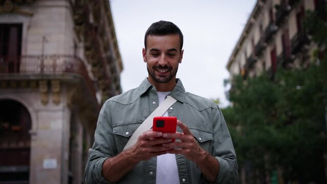 Young Caucasian attractive man walking looking happy mobile phone on street in European city of Barcelona. Joyful male tourist strolling using red cell enjoys typing outdoor travel tourism experience