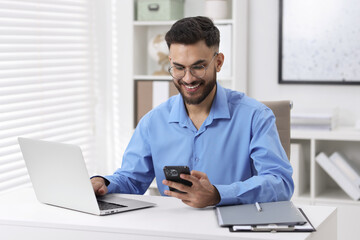 Happy young man using smartphone while working with laptop at white table in office