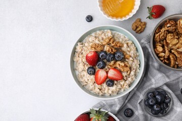 Tasty oatmeal with strawberries, blueberries and walnuts surrounded by ingredients on grey table, flat lay. Space for text
