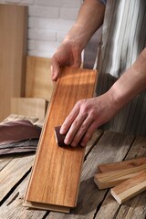 Man polishing wooden plank with sandpaper at table indoors, closeup - obrazy, fototapety, plakaty