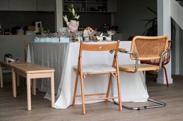 A table covered with long white tablecloth in industrial loft kitchen is decorated with a pink handmade vase with lilies, blue and gold boxes, champagne glasses with raspberries, surrounded by chairs.