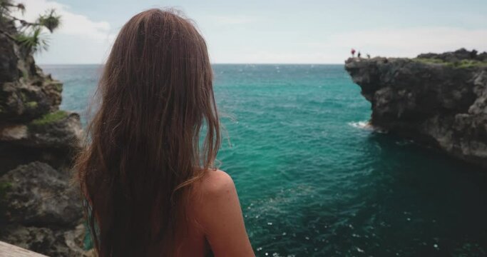 A woman stands confidently on the edge of a cliff overlooking the vast ocean on Wild Sumba Island in Indonesia. The Weekurri Lagoon Salt Lake sparkles in the distance.