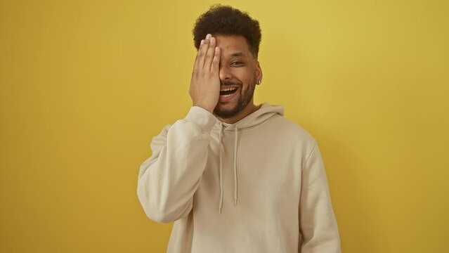 Confident african american man in fashion-forward hoodie, teasingly covering eye with his hand, beaming a hearty smile over isolated yellow backdrop.