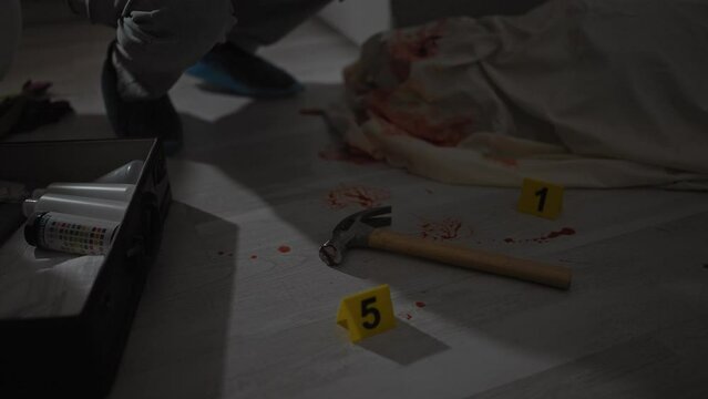 An investigator examines a bloody hammer at a dimly-lit indoor crime scene with evidence markers.