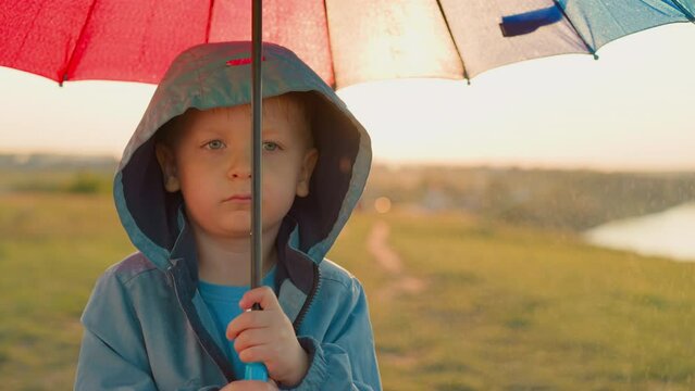 Upset kid with umbrella at rainfall on riverbank. Thoughtful little boy in bad mood stands alone in green field on rainy day. Child in discomfortable place