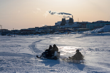 People on a snowmobile with sleds ride along the bank of a frozen river. Winter arctic industrial landscape. industry and ecology in the Arctic. Everyday life in the Far North of Russia. Chukotka.