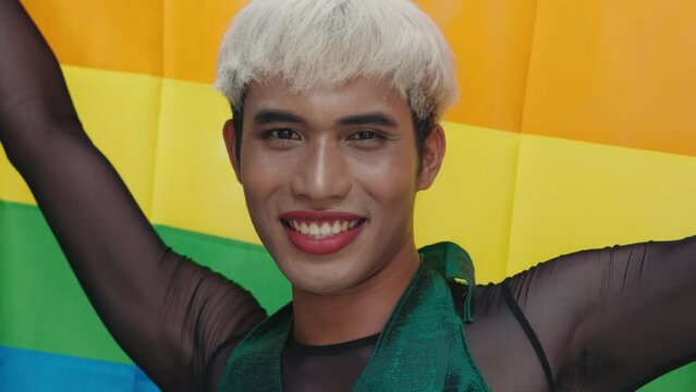 Young Asian Transgender Man Standing and Showing LGBT Community Flag to Camera. Nicely Dressed Man with Painted Lips and Makeup Outside Pose with Rainbow Flag. Diversity LGBT Concept