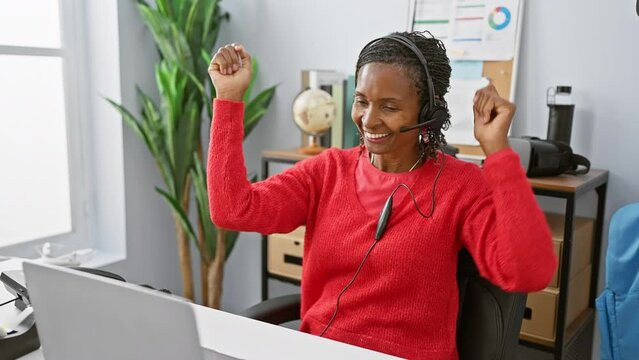 Excited woman celebrating success at workplace with computer, headphones, office, joy, technology, professional, african, mature, desktop, interior