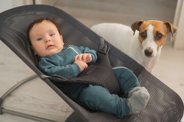 A dog sits next to a cute three-month-old boy dressed in a blue overalls in a baby lounger.