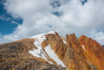 Snow-capped mountain ridge with cornice and red sharp rocks on top in sunlight. Snow cap and shiny pointy peak of gold color. Snow dome and colorful rocky peaked top. Sunny cloudy high mountains view.