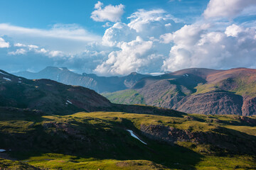 Awesome alpine landscape with sunlit green hills and multicolor mountains under huge lush clouds in...