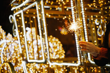 Woman holding sparkler night while celebrating Christmas outside. Dressed in a fur coat and a red headband. Blurred christmas decorations in the background. Selective focus