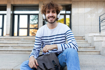 Happy young university student sitting on stairs in campus wearing headphones looking at camera.