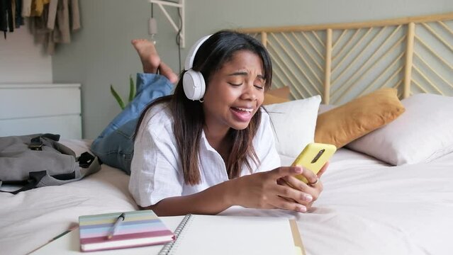 4k resolution video of female Black teen college student using phone and headphones listening to music lying down on bed