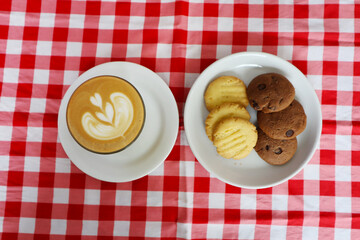 Choco chip cookies and latte coffee on a white plate with a checkered tablecloth seen above