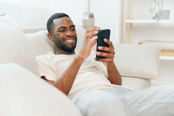 Young African American man sitting on a black sofa in his modern apartment, happily typing a...