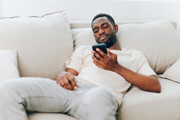 Happy African American man sitting on a black sofa, holding a mobile phone, typing a message He is...