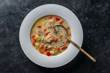 Fresh creamy salmon fish soup with potato, carrots, tomato, peppers and onions in a white ceramic plate on a black background, closeup, top view. A delicious dinner consists of fish soup with salmon - 778650674