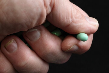 Prescription medicine (drugs) misuse, abuse. Green capsules held in closed hand. One capsule clasped between thumb and index finger. Black background. Copy space.