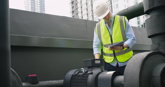 A Engineer man looking inspecting maintenance insulated pipelines valve pump control on the roof at an industrial site. He is wearing a hard hat and safety vest