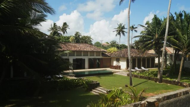 Approaching drone shoots video of luxury retro villa on oceanfront island with access to white sandy beach. Side view of cottages with swimming pools and palm trees in resort area of Sri Lanka.