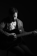 Black and white studio portrait of a caucasian man in his 40s playing an acoustic guitar. He is wearing a jean jacket and white shirt underneath. He is attractive and has brown hair and a beard. 