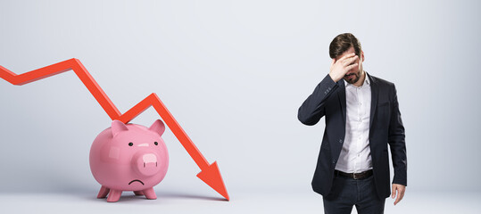 Stressed young businessman with falling red chart arrow with piggy bank on white background. Economic recession, losing savings concept.