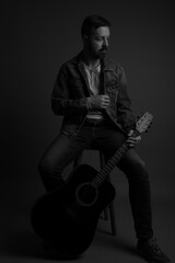 Fototapeta premium Black and white studio portrait of a handsome caucasian man sitting on a stool and holding an acoustic guitar. He is wearing running shoes, jeans, jean jacket and a white shirt underneath. 