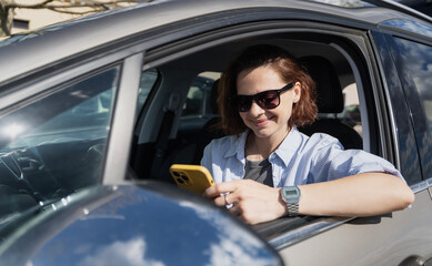 Smiling Caucasian woman in sunglasses sitting in a car in the parking lot driver's seat looking at the smartphone - 778726293