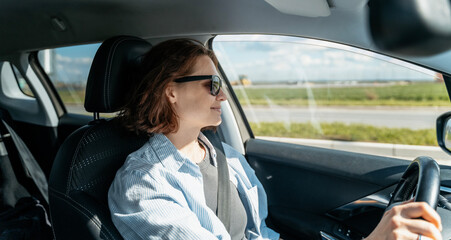 Young Caucasian woman in sunglasses driving a car in the countryside on a summer day - obrazy, fototapety, plakaty