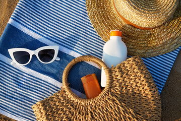 Beach essentials laid on striped towel at golden hour. Woven straw, oversized white sunglasses, sun cream bottles in natural tote bag imply summertime skin care. Holiday scene by sea shore.