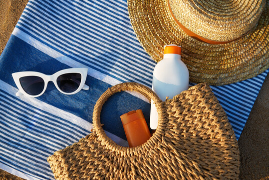 Beach essentials laid on striped towel at golden hour. Woven straw, oversized white sunglasses, sun cream bottles in natural tote bag imply summertime skin care. Holiday scene by sea shore.