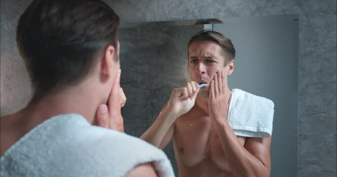 Attractive man brushes teeth in bathroom in morning. While brushing feels sharp pain in tooth and stops. Holding his aching tooth man looks into mirror with toothbrush in his hand. Sick tooth concept.