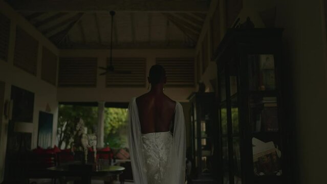 Elegant androgynous person of color walks model gait around villa and shows his white dress, perfect skin. Exposure shooting of non-binary black man posing outside house on tropical island.
