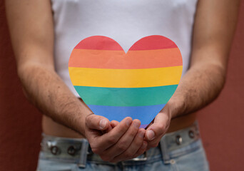 An unrecognizable gay male is holding a rainbow heat symbol with both hands beside his body to...