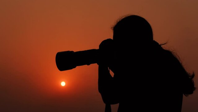 A young photographer is taking pictures with a professional camera in the color of a golden summer sunset sky.