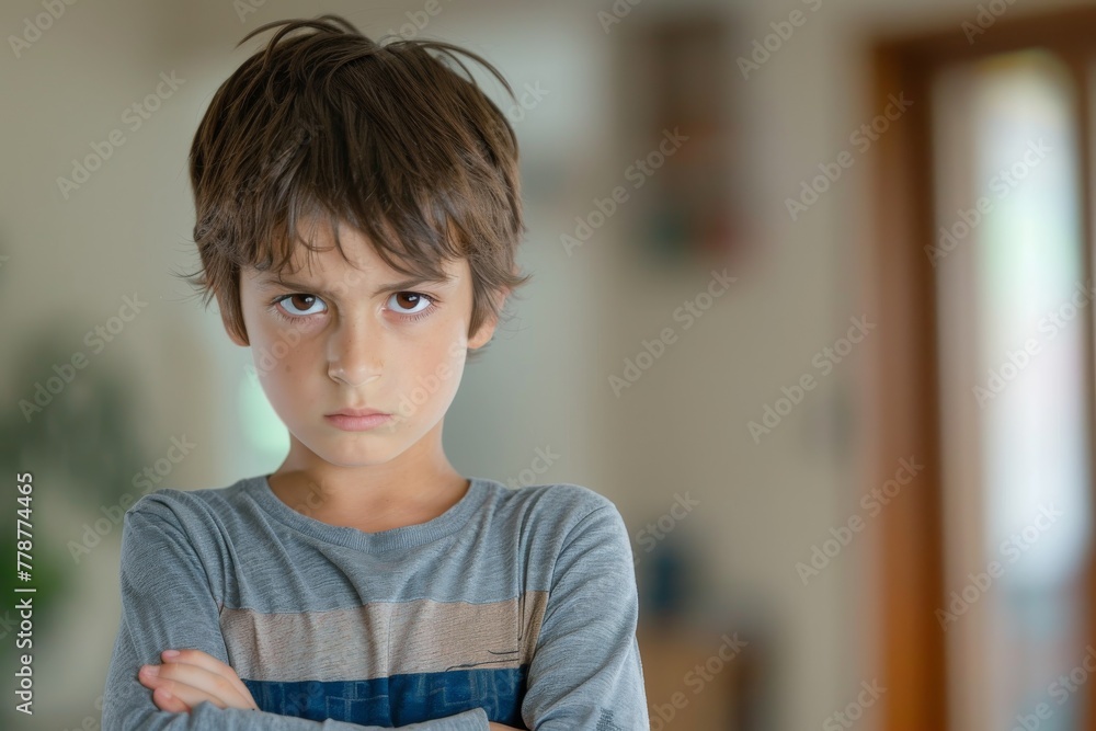 Wall mural Close-up of a discontent young boy arms crossed, standing inside a room with a blurred backdrop