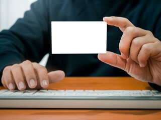 Businessman showing blank white plain namecard while working with keyboard computer on wooden table. Mockup business name card, horizontal style holding by hand of man in black shirt.