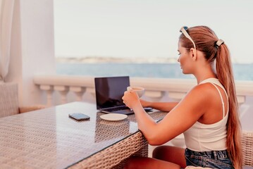 woman is sitting at a table with a laptop and a cup of coffee. She is wearing sunglasses and a ponytail. The scene suggests a relaxed and leisurely atmosphere