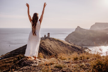 A woman in a white dress stands on a rocky hill overlooking the ocean. She is smiling and she is...
