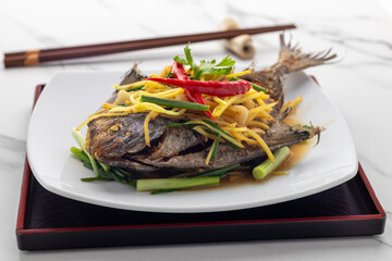 Fried whole black pomfret with shredded ginger and soybean paste sauce, a simple delicious Thai dish presents that Thai food also does reflect Chinese cuisine, served on a white serving plate.