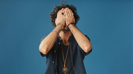 Annoyed man with curly hair, dressed in blue shirt, dissatisfied covers his face with his palms...