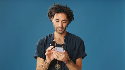 Man with curly hair, dressed in blue shirt, using mobile phone, typing massages isolated on blue...