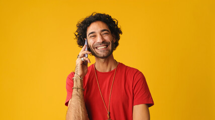 Smiling man with curly hair, dressed in red T-shirt,  talking on mobile phone isolated on yellow...