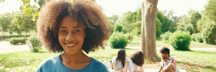 Portrait of young smiling woman with curly hair wearing blue t-shirt posing for the camera in the park, Panorama. Picnic on summer day outdoors her friends sitting in distance blurred on background
